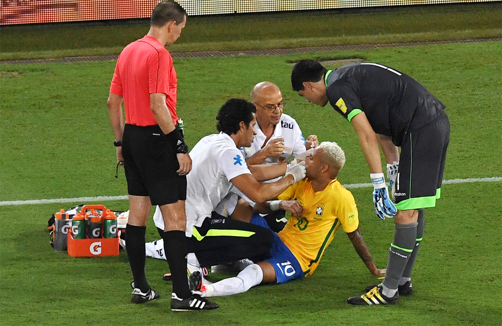 Tiền đạo Neymar (giữa, ngồi dưới đất) của đội tuyển Brazil bị thương đổ máu ở đầu trong khi tranh chấp bóng được các nhân viên y tế săn sóc ngay trên sân cỏ trong trận đấu vòng loại World Cup 2018 giữa Brazil và Bolivia diễn ra tại Natal, Brazil ngày 6 tháng 10, 2016. (Hình: Getty Images)