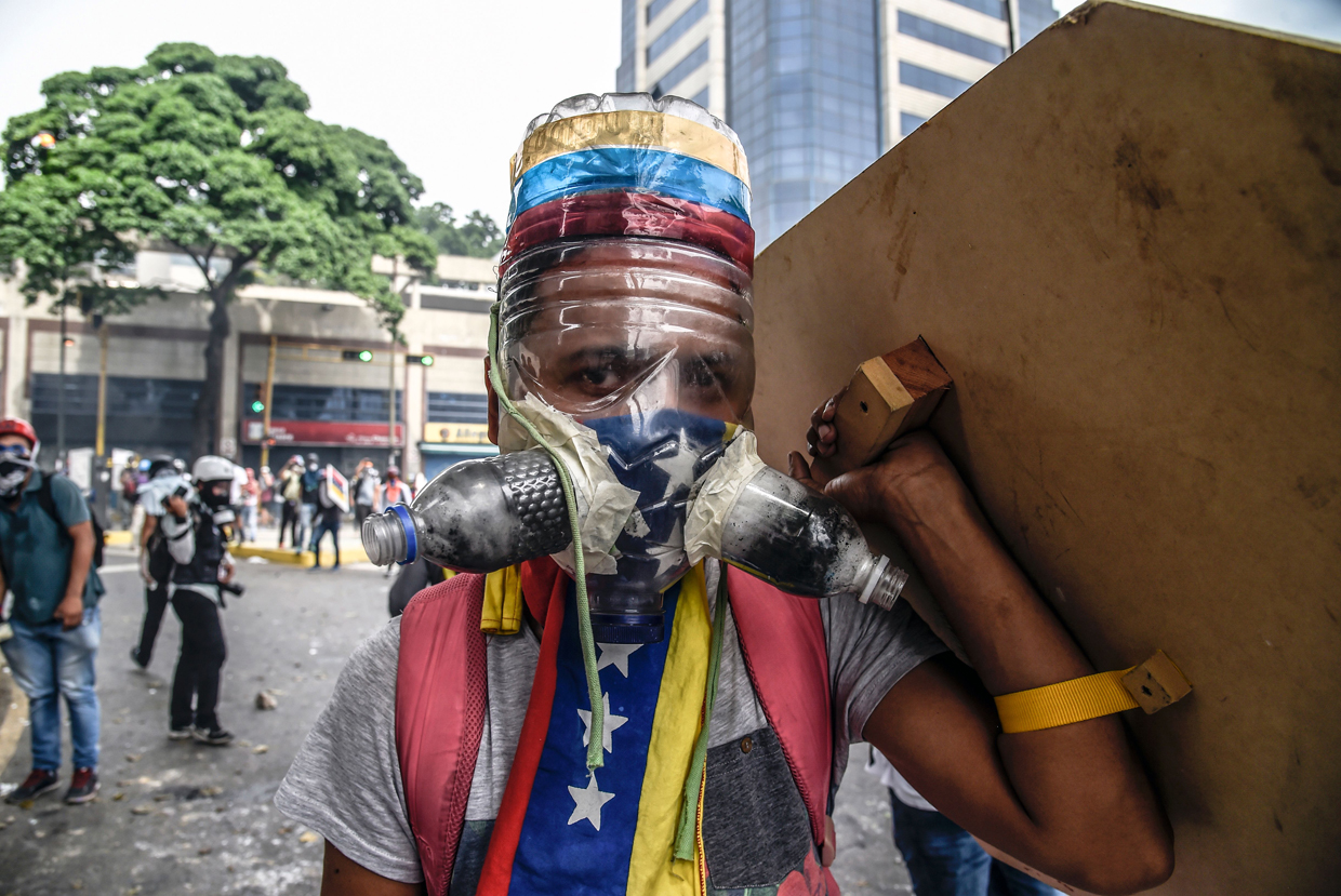 TOPSHOT - A Venezuelan opposition activist wearing a homemade gas mask takes cover behind a makeshift shield as clashes erupt with riot police during a protest against President Nicolas Maduro, in Caracas on May 8, 2017. Venezuela's opposition mobilized Monday in fresh street protests against President Nicolas Maduro's efforts to reform the constitution in a deadly political crisis. Supporters of the opposition Democratic Unity Roundtable (MUD) gathered in eastern Caracas to march to the education ministry under the slogan "No to the dictatorship." / AFP PHOTO / JUAN BARRETO (Photo credit should read JUAN BARRETO/AFP/Getty Images)